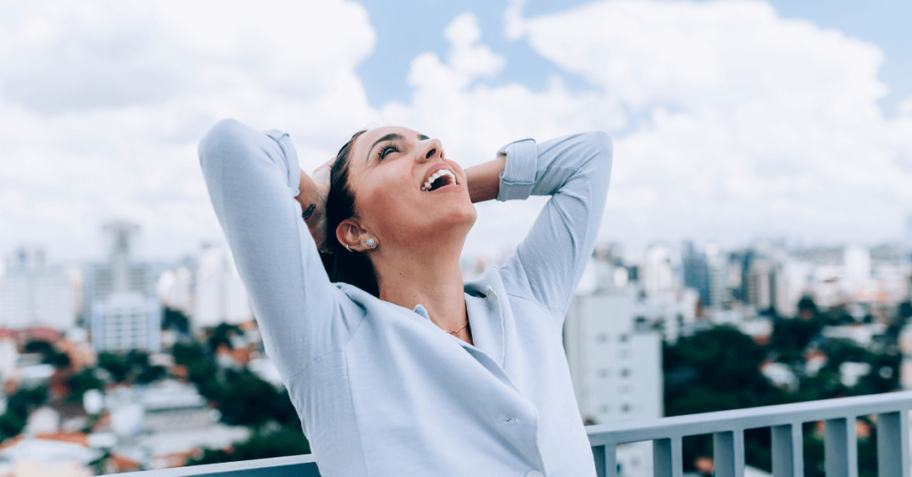 Imagem de uma mulher sentada em uma cadeira, em ambiente urbano aberto, olhando para o céu, com as mãos na cabeça, e uma expressão de alívio, contemplação e alegria. Imagem destacada do site louvare.com, referente ao post: Como encontrar ajuda divina e alívio em momentos de angústia