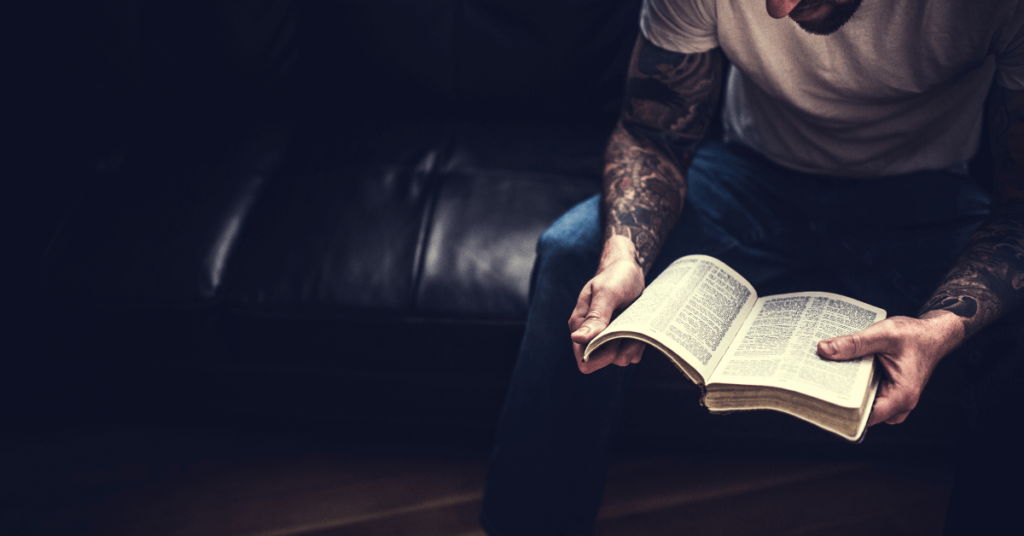 Imagem de um homem com tatuagens nos dois braços, sentado e lendo a Bíblia Sagrada em busca de respostas, conselhos e ensinamentos divinos. Imagem destacada do site louvare.com, referente ao post: O que a Bíblia diz sobre tatuagem?
