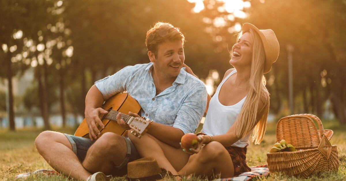 Imagem de um casal feliz e alegre, rindo enquanto tocam violão, sentados em meio a natureza. Imagem destacada do site louvare.com, referente ao post: Oração pela construção de relacionamentos saudáveis e significativos