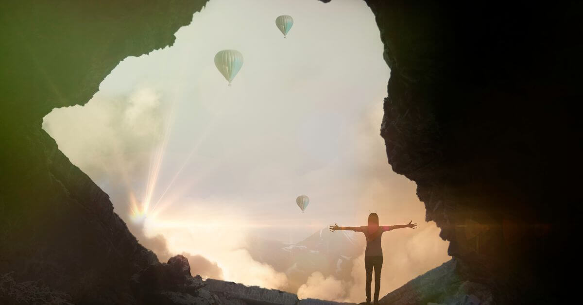 Imagem de uma mulher, com os braços abertos, na entrada de uma caverna, em um lindo ambiente natural. Ao fundo encontra-se um céu lindo repleto de balões grandes. Imagem destacada do site louvare.com, referente ao post: Reflexão: Versículo Lucas 1:37 – Crescimento e Esperança