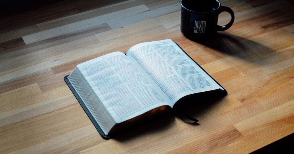 Imagem de uma Bíblia aberta sobre uma mesa, ao lado de uma caneca preta. Imagem destacada do site louvare.com, referente ao post: O que a Bíblia diz sobre São João?