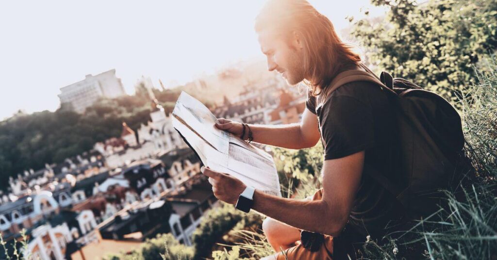 Imagem de um jovem viajante, sentado em uma colina, lendo um mapa ao ar livre com uma cidade ao fundo, representando a busca por direção e propósito na vida. Essa imagem destaca o tema de estar no lugar certo e na hora certa, conforme discutido no site Louvare.com. Imagem destacada do post: "O que a Bíblia diz sobre estar no lugar certo, na hora certa?".